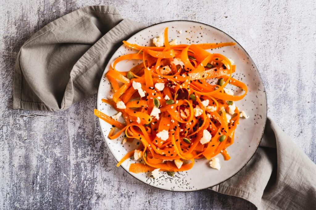 Carrot ribbon salad with feta and toasted seeds on a speckled plate, styled with a grey napkin.