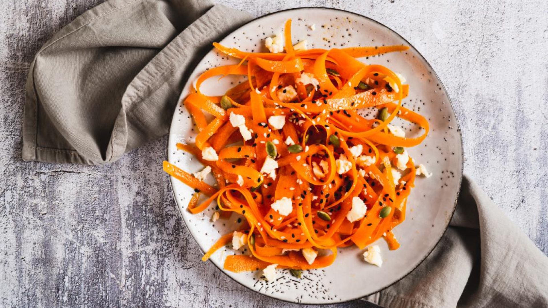 Carrot ribbon salad with feta and toasted seeds on a speckled plate, styled with a grey napkin.